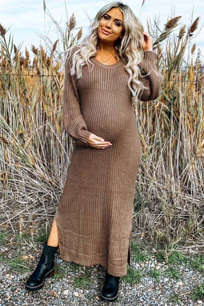 woman wearing a brown ribbed maternity sweater dress and holding her baby bump in a field with tall grass