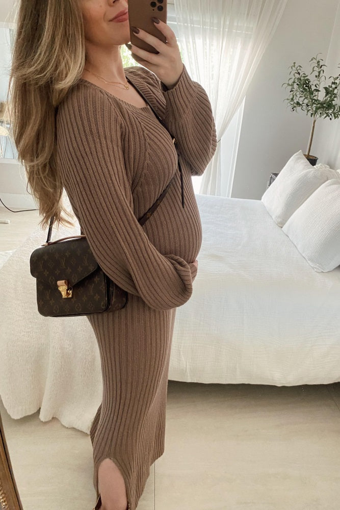 woman wearing a brown ribbed maternity dress, showing off her baby bump and holding a handbag, in a bright room