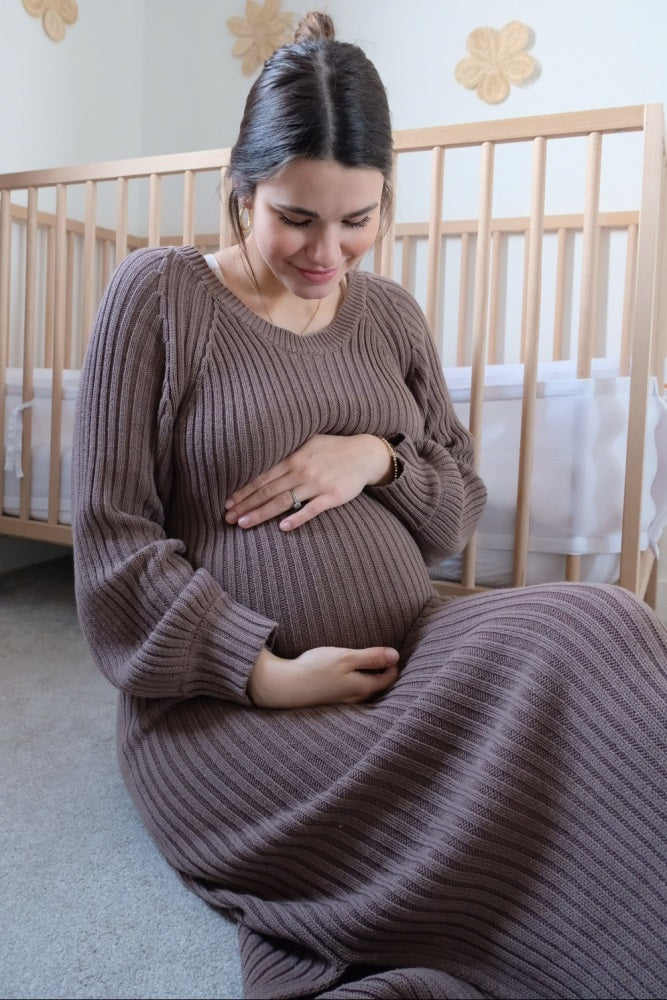 woman wearing a ribbed brown maternity dress, sitting and holding her baby bump in a cozy nursery setting