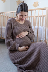 woman wearing a ribbed brown maternity dress, sitting and holding her baby bump in a cozy nursery setting