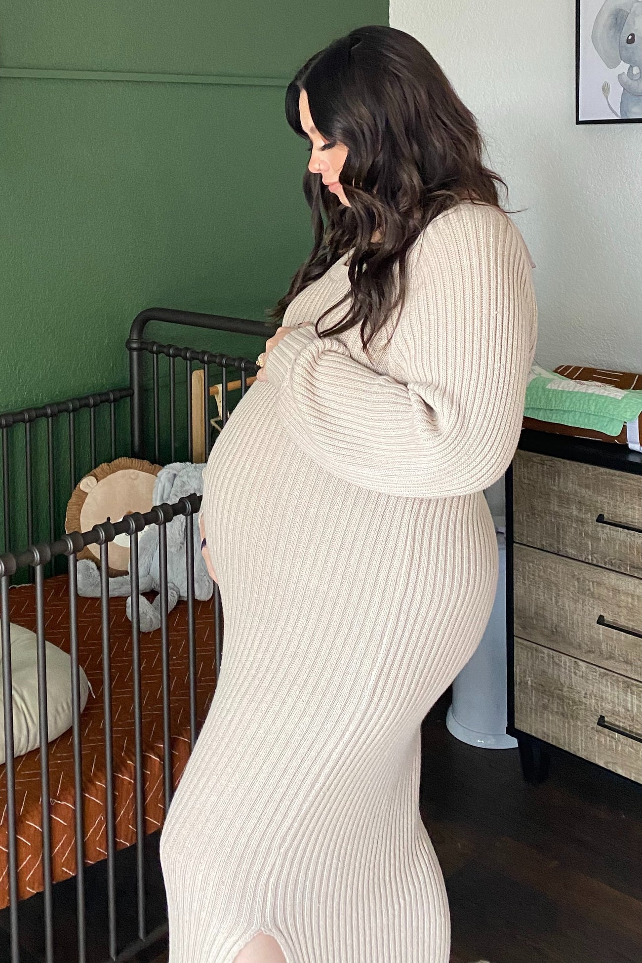 woman wearing a beige ribbed maternity dress highlighting her baby bump while standing beside a crib