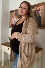 smiling pregnant woman in a tan cardigan and black shirt holding her baby bump in a home setting