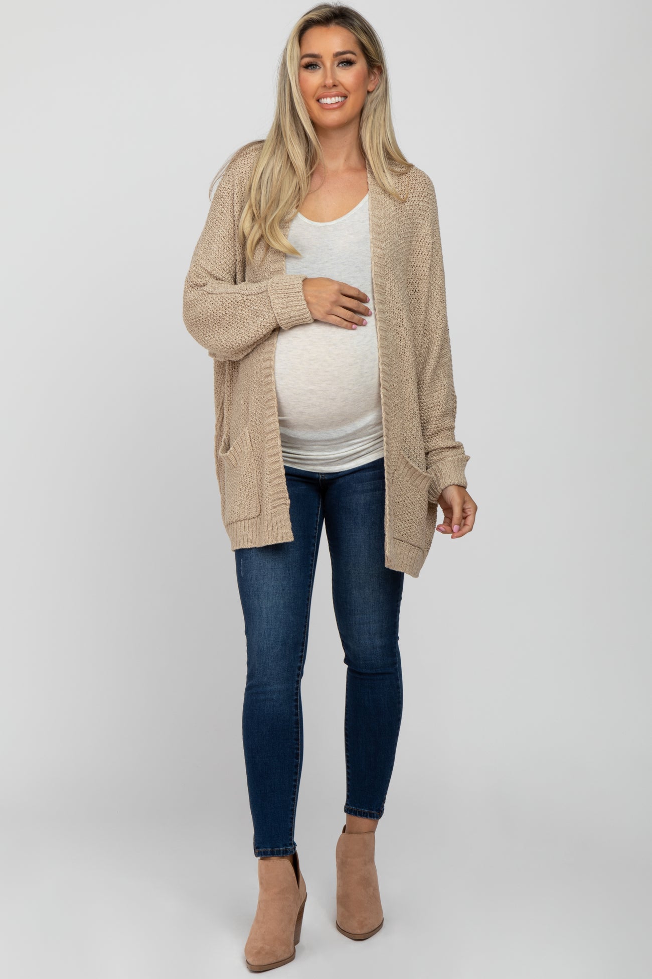 woman wearing a beige maternity cardigan over a light gray top and showing her baby bump with skinny jeans and ankle boots