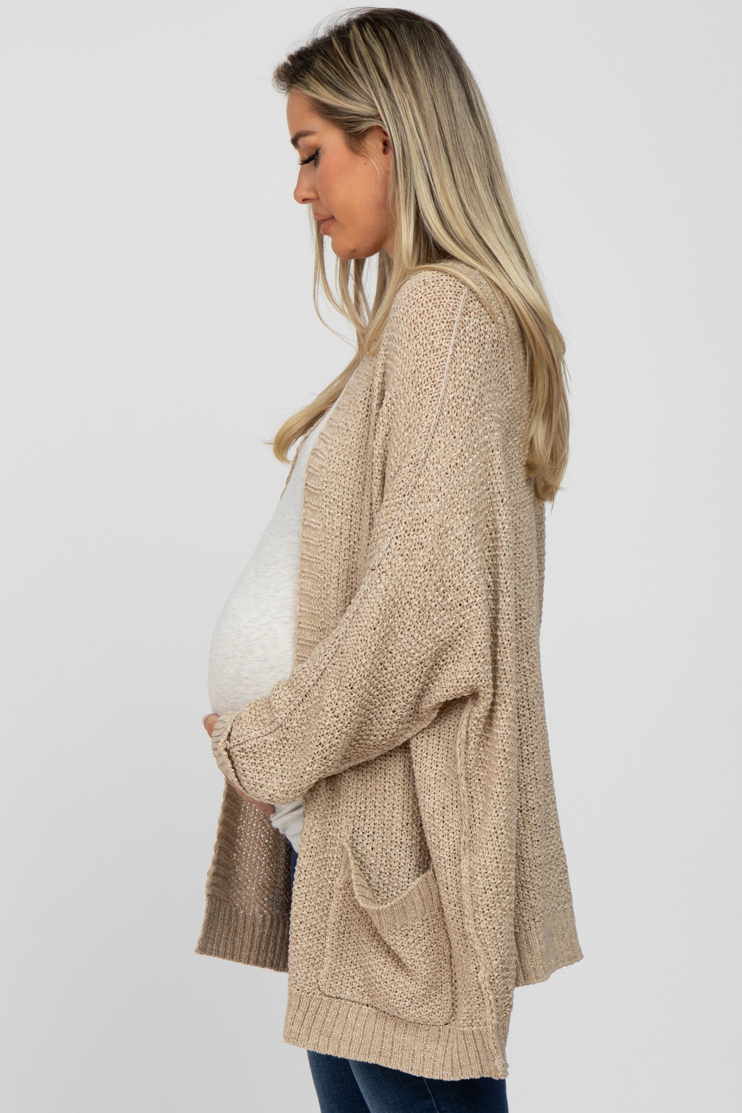 woman wearing a light beige open-knit maternity cardigan and holding her baby bump