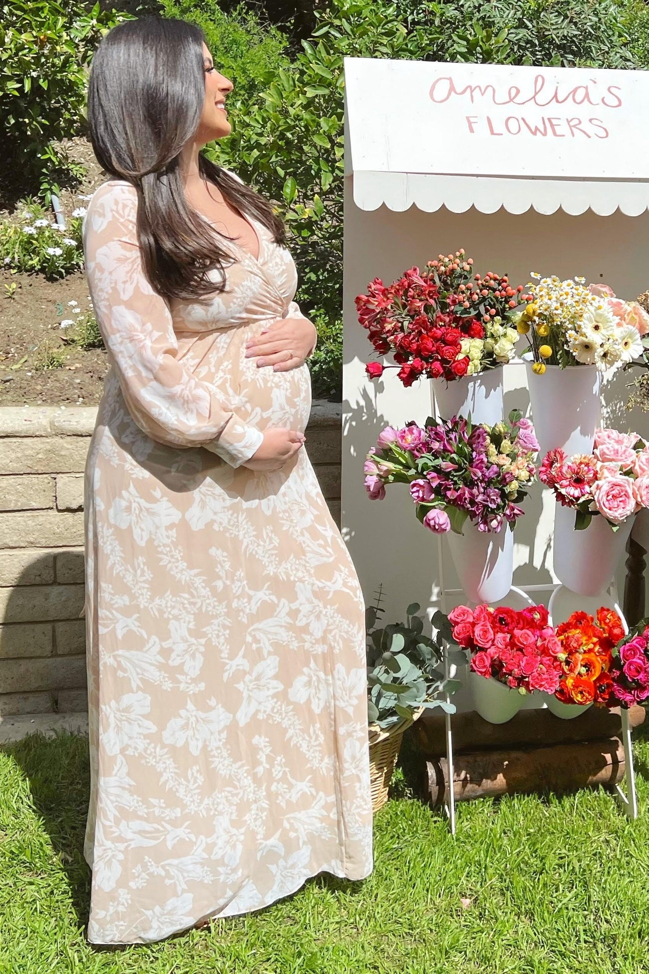 woman wearing a beige floral long sleeve maternity dress and holding her baby bump outdoors by a flower stand