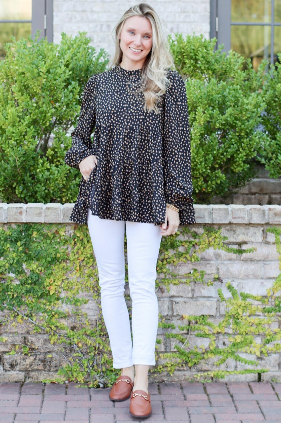 woman wearing a black polka dot long sleeve peplum top and white skinny jeans, standing outdoors with greenery in background