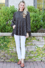 woman wearing a black polka dot long sleeve peplum top and white skinny jeans, standing outdoors with greenery in background