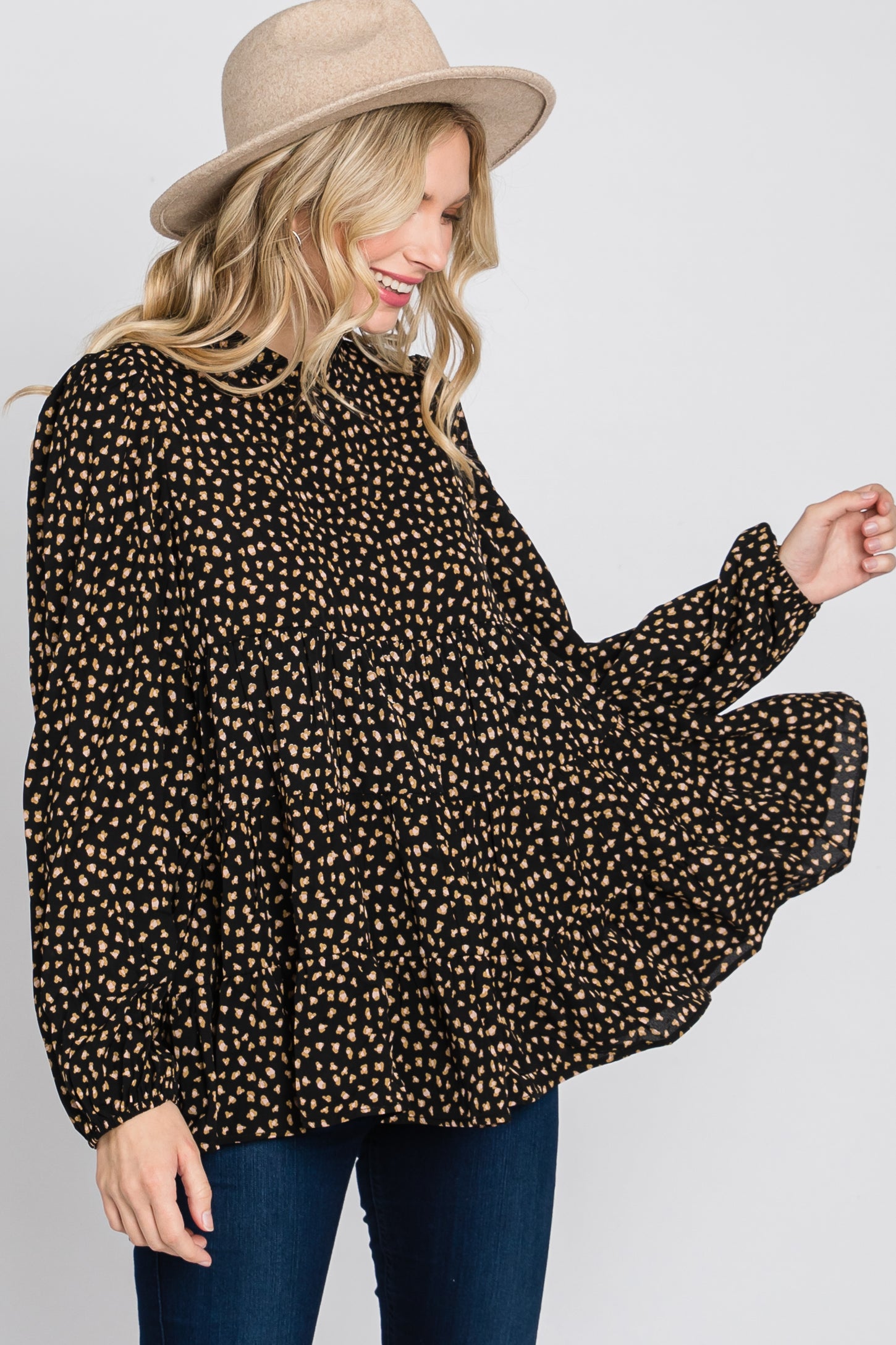 woman wearing a black and tan polka dot button-up shirt with a flowy silhouette and long sleeves, smiling while wearing a hat