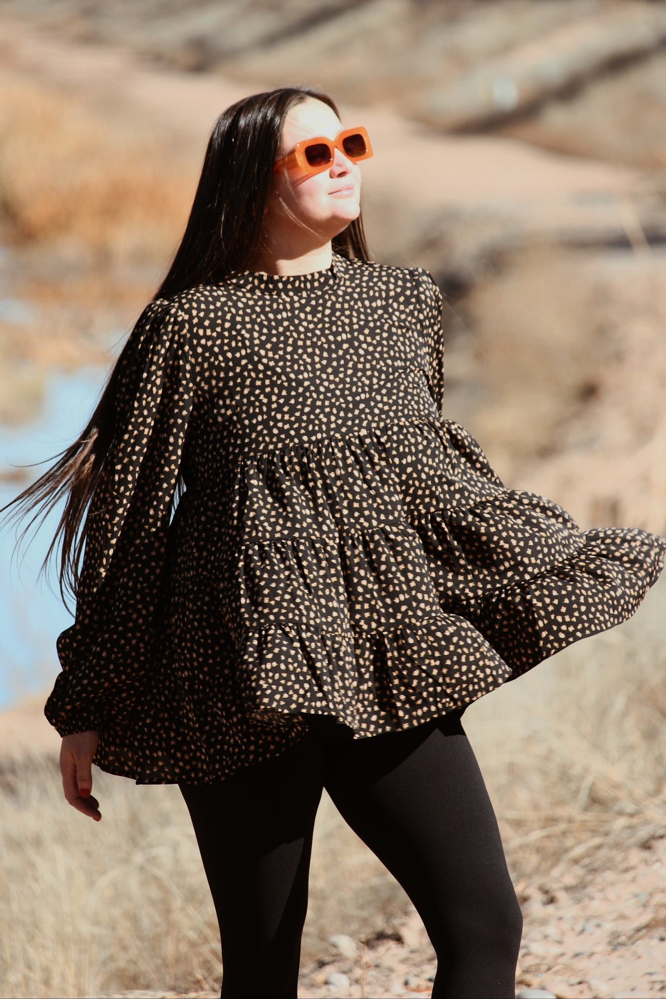 woman wearing a black and tan polka dot blouse with puff sleeves and a flared hem, paired with black leggings and sunglasses
