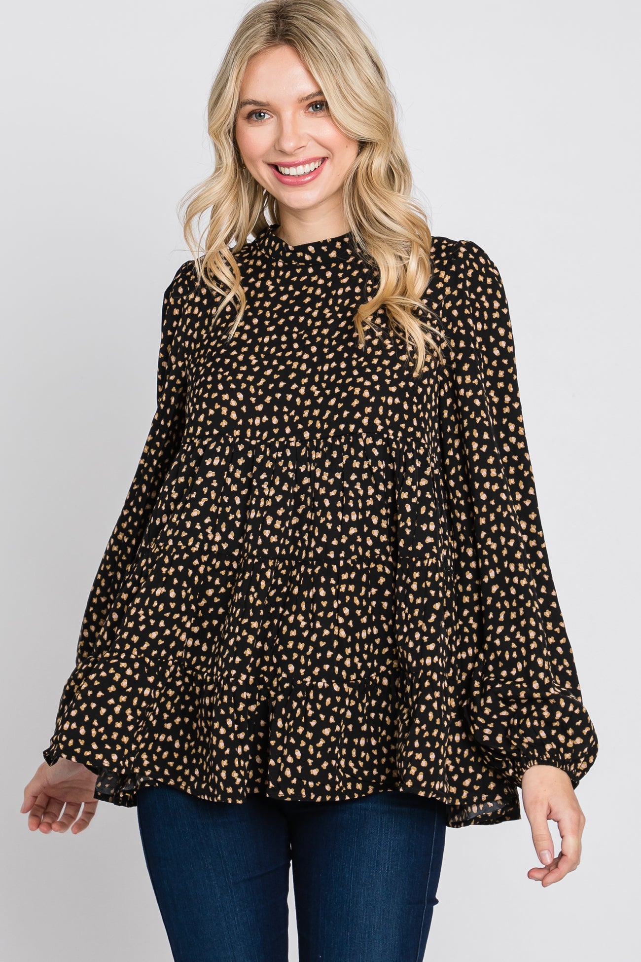 woman wearing a black blouse with tan polka dots and bell sleeves, smiling and posing against a plain background