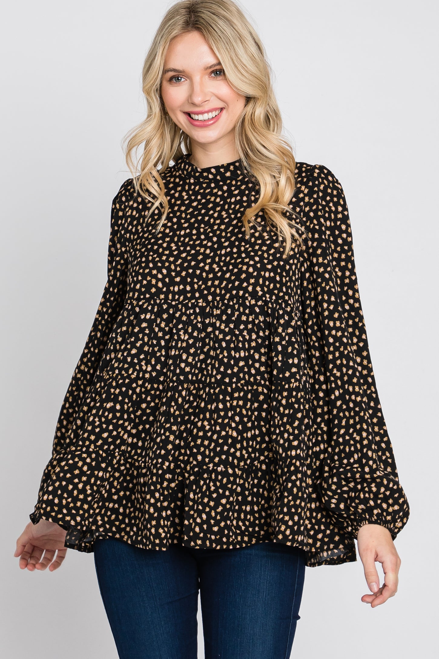 woman wearing a black blouse with tan polka dots and bell sleeves, smiling and posing against a plain background