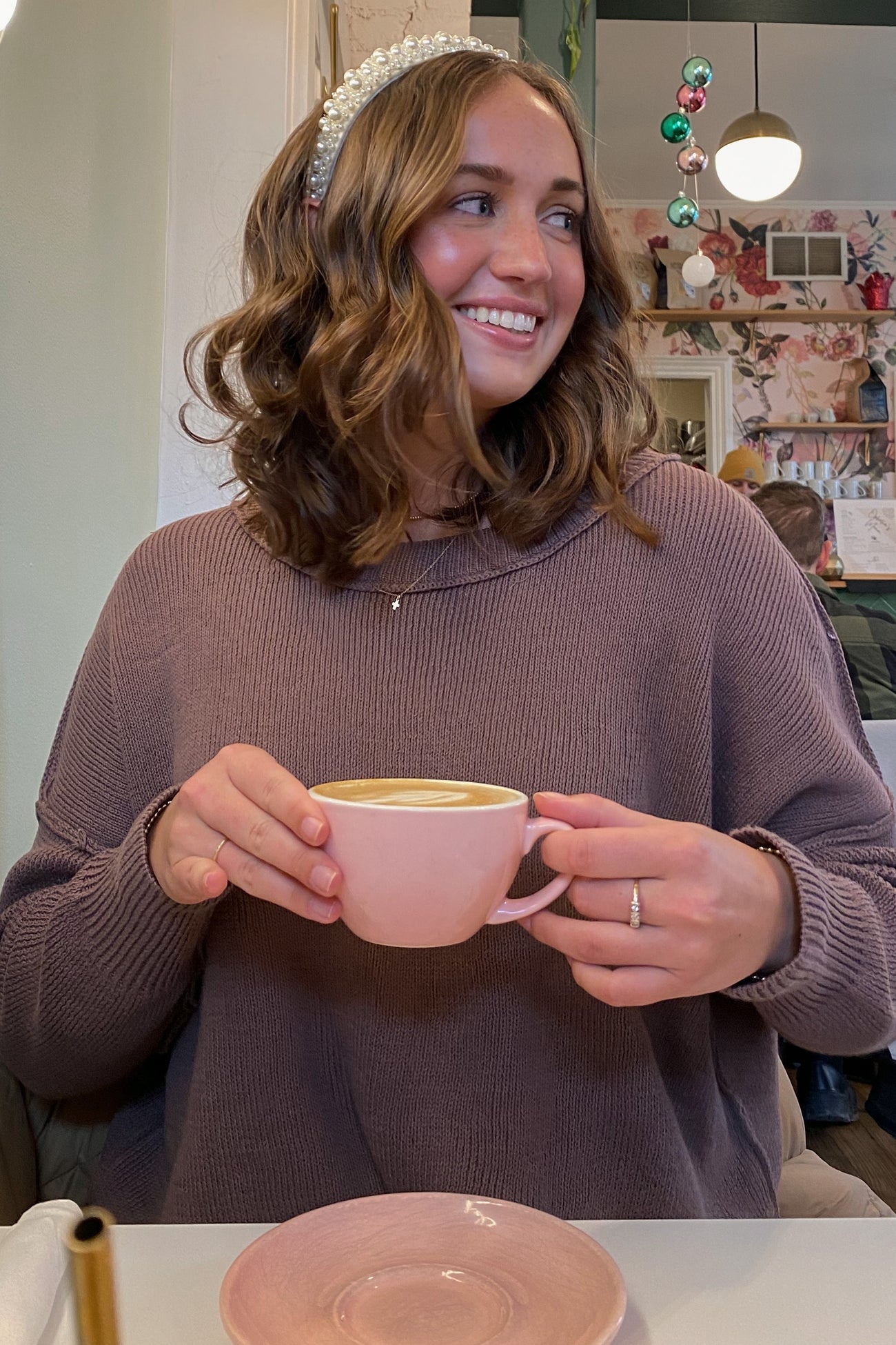 woman wearing a mauve sweater and holding a pink coffee cup, smiling with a decorative headband in a cafe setting
