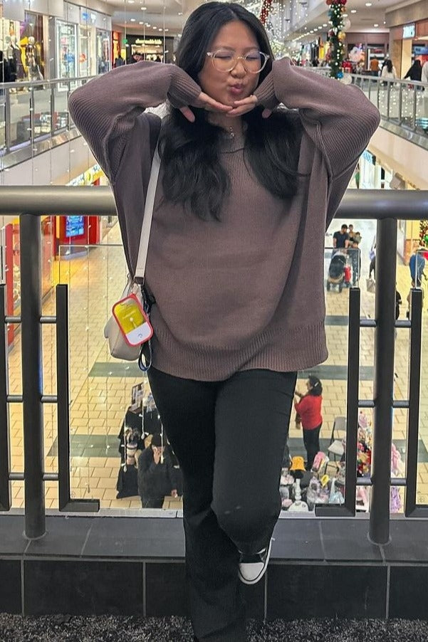 woman wearing a oversized brown sweater and black pants, posing with hands under her chin in a shopping mall