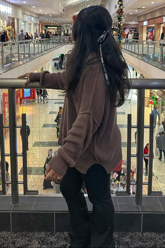 woman wearing a brown oversized sweater and black flared pants, standing with her back to the camera in a shopping mall