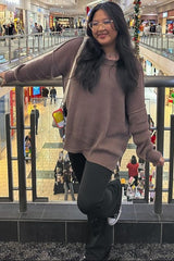 woman wearing a brown oversized sweater and black flared pants, posing with one hand on a railing in a shopping mall