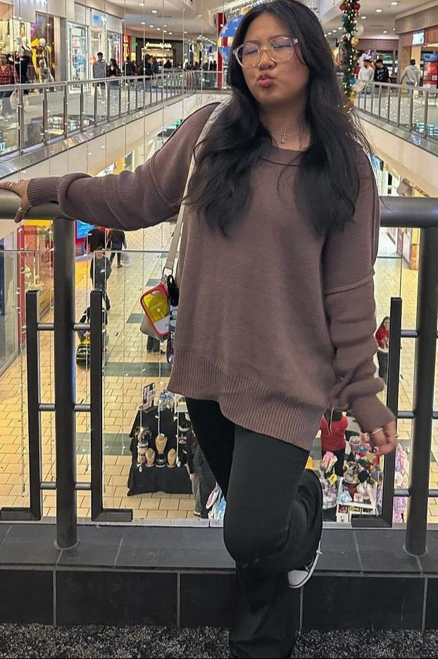 woman wearing a brown oversized sweater and black pants, posing casually by a railing in a shopping mall
