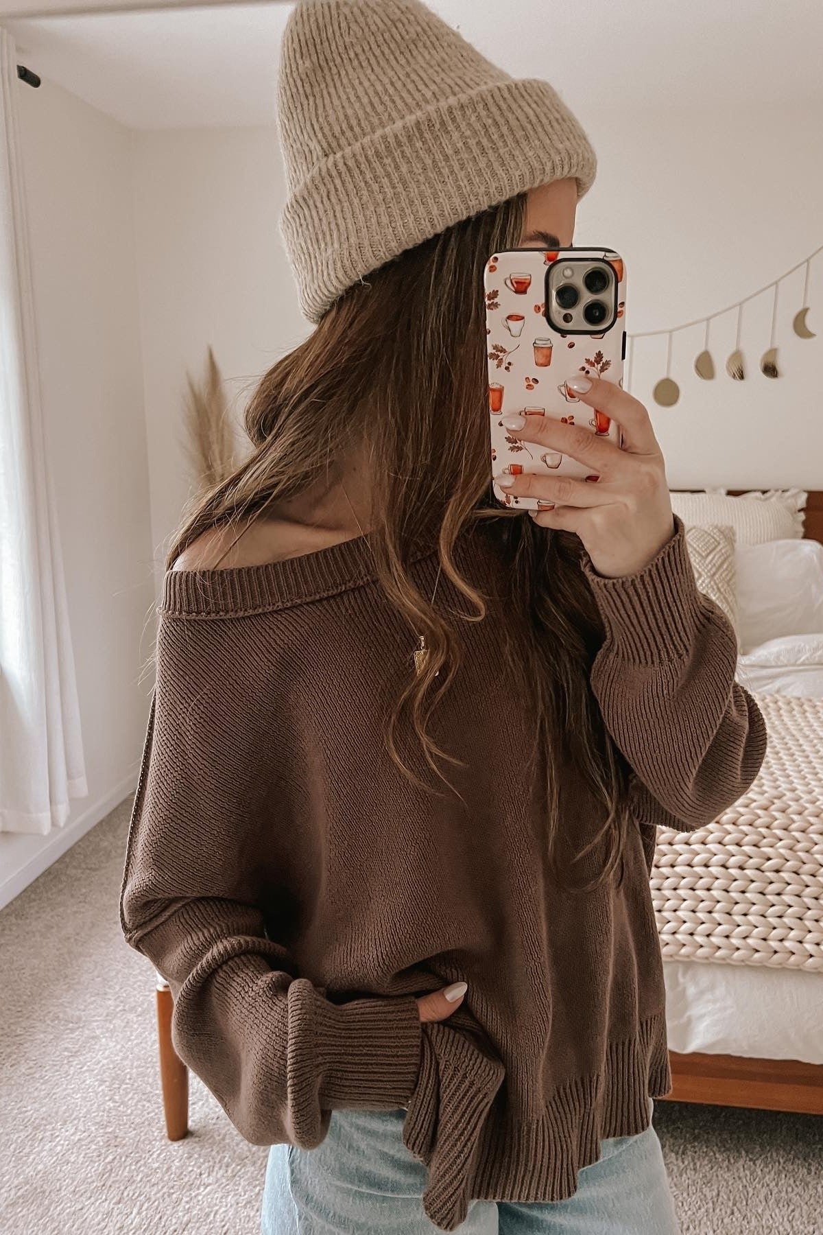 woman wearing a brown off-shoulder sweater and light blue jeans, holding a phone with a patterned case
