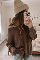 woman wearing a brown off-shoulder sweater and light blue jeans, holding a phone with a patterned case