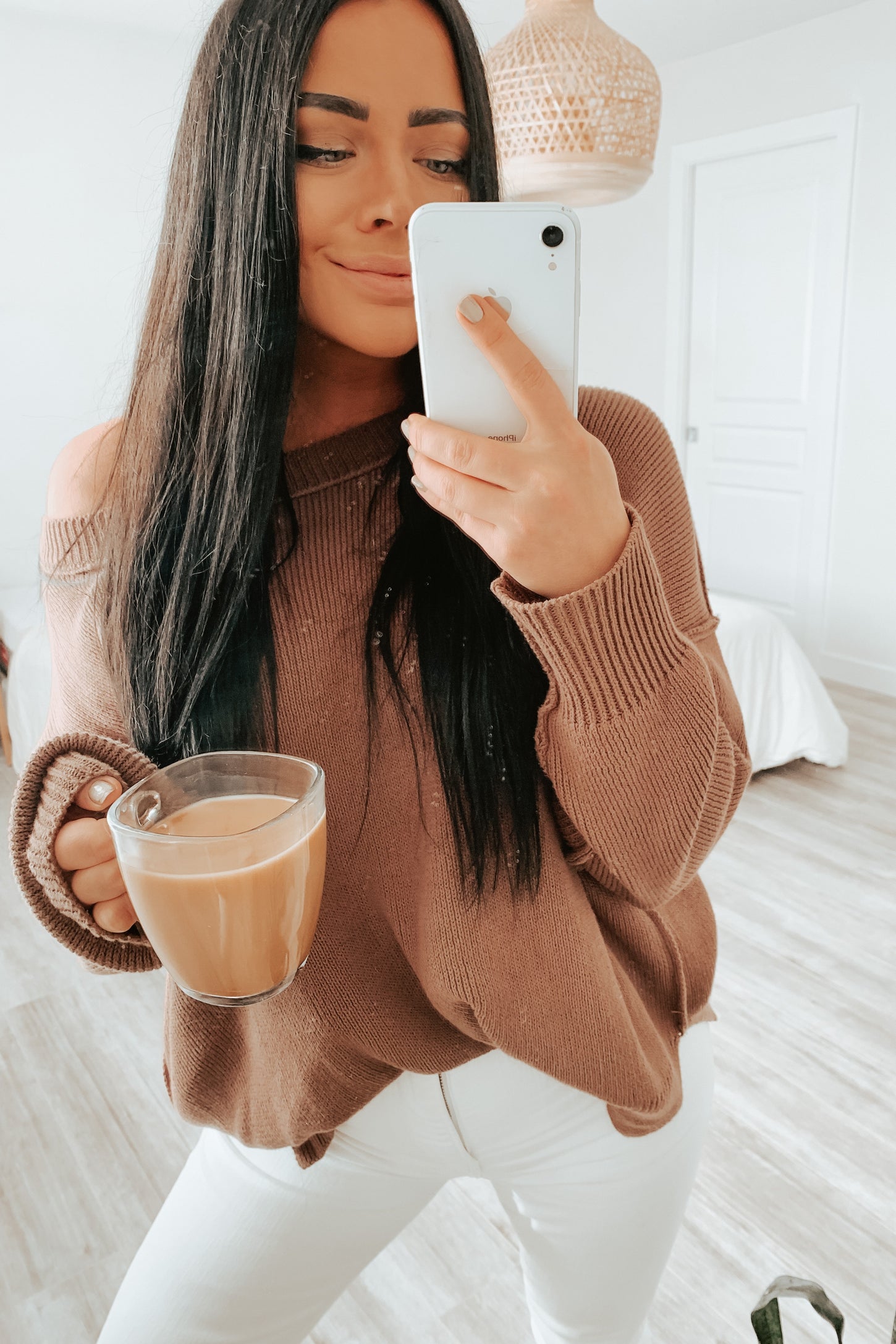 woman in brown knit sweater and white jeans taking a mirror selfie while holding a glass of coffee