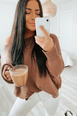 woman in brown knit sweater and white jeans taking a mirror selfie while holding a glass of coffee
