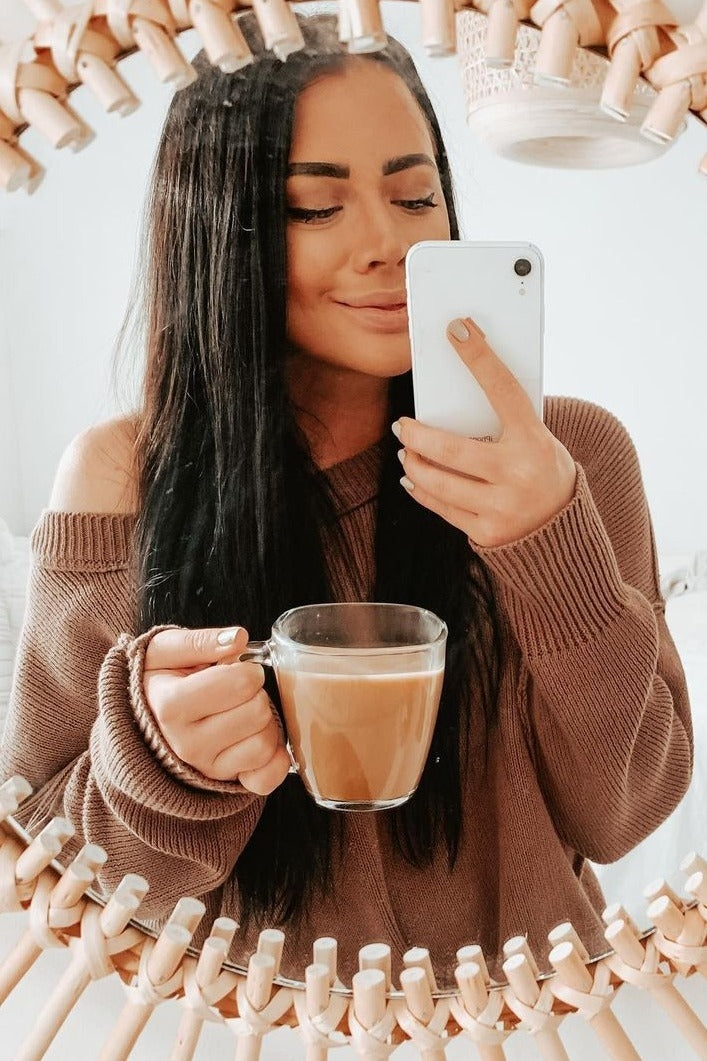 woman wearing a cozy brown off-shoulder sweater holding a cup of coffee while looking at her reflection in a mirror