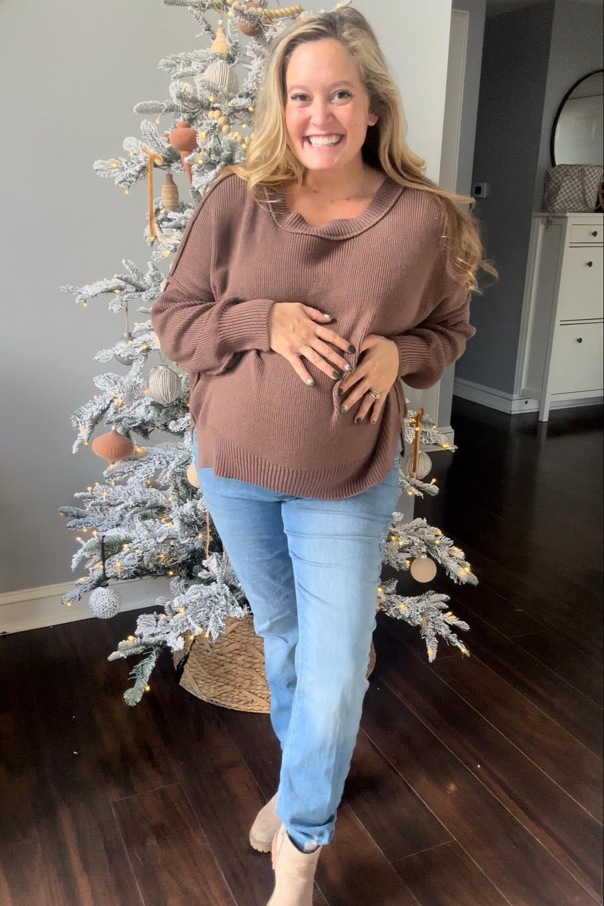 woman wearing a brown oversized maternity sweater and light blue jeans, showing her baby bump, smiling in front of a decorated Christmas tree