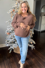 woman wearing a brown oversized maternity sweater and light blue jeans, showing her baby bump, smiling in front of a decorated Christmas tree