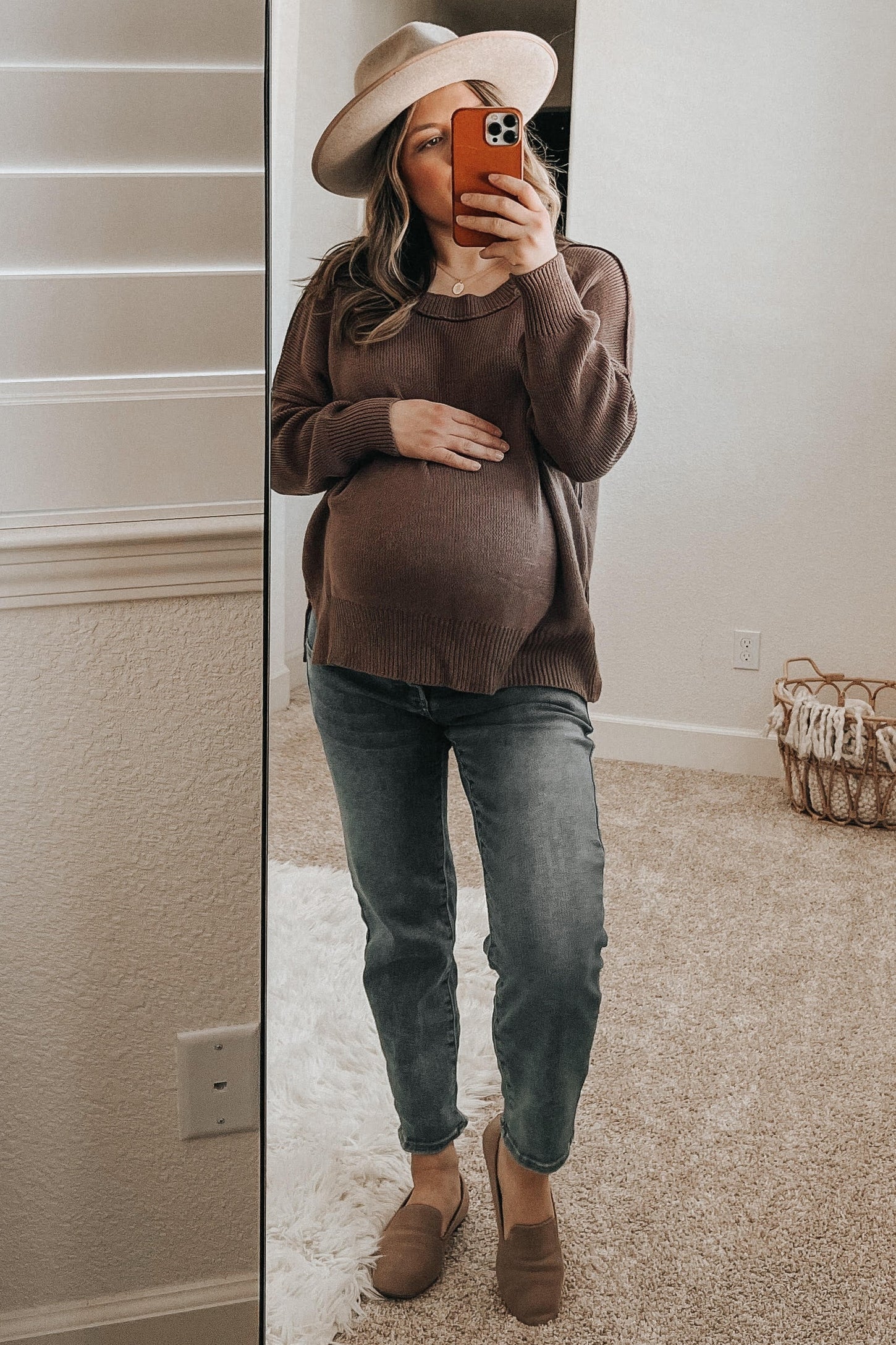 woman wearing a brown oversized maternity sweater and light blue jeans, holding her baby bump in front of a mirror