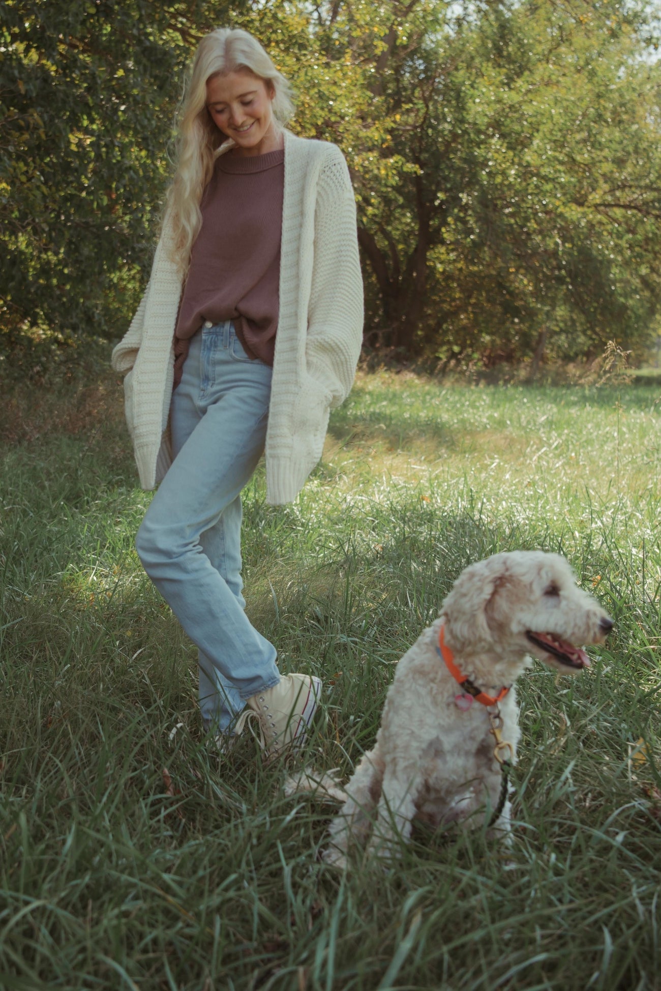 woman wearing a cozy cream cardigan over a purple top and light blue jeans, standing in a grassy area with a dog