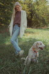 woman wearing a cozy cream cardigan over a purple top and light blue jeans, standing in a grassy area with a dog