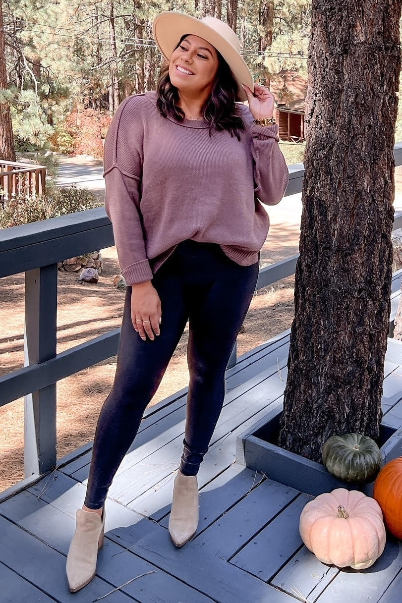woman wearing a mauve oversized sweater and black leggings, standing outside with pumpkins and trees in the background