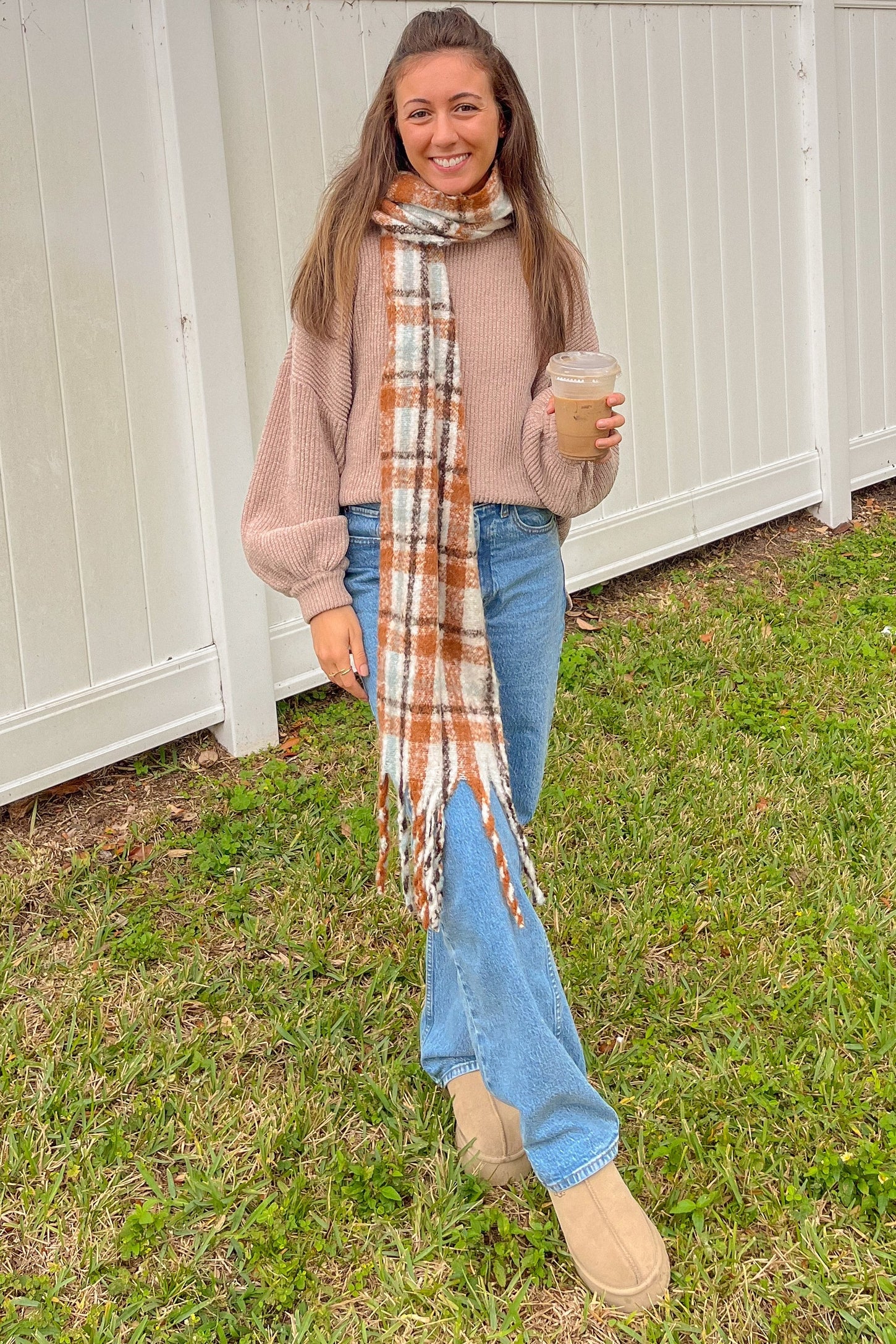 woman wearing a beige oversized sweater, light blue jeans, brown suede boots, and a plaid scarf, holding a drink