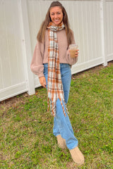 woman wearing a beige oversized sweater, light blue jeans, brown suede boots, and a plaid scarf, holding a drink