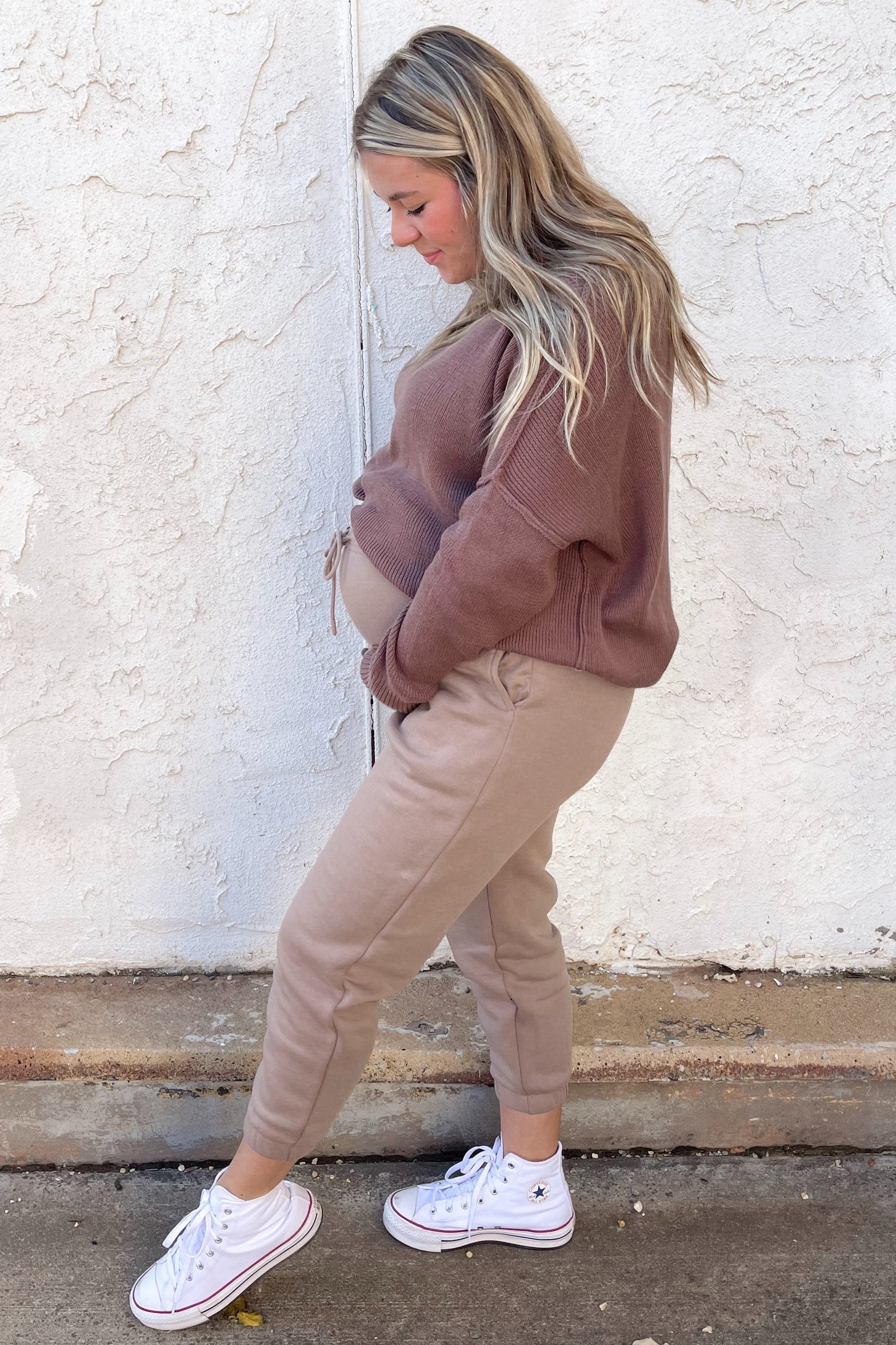 woman wearing a brown maternity sweater and beige jogger pants, holding her baby bump while standing sideways