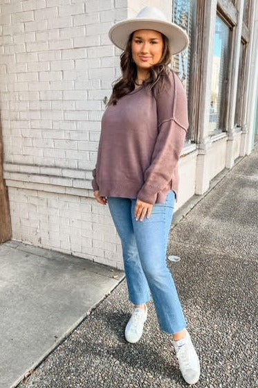 woman wearing a mauve oversized sweater, light blue jeans, and white sneakers, standing outside with a wide-brim hat