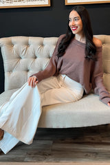 woman wearing a brown off-shoulder sweater and white wide-leg trousers, sitting on a tufted sofa with a smile