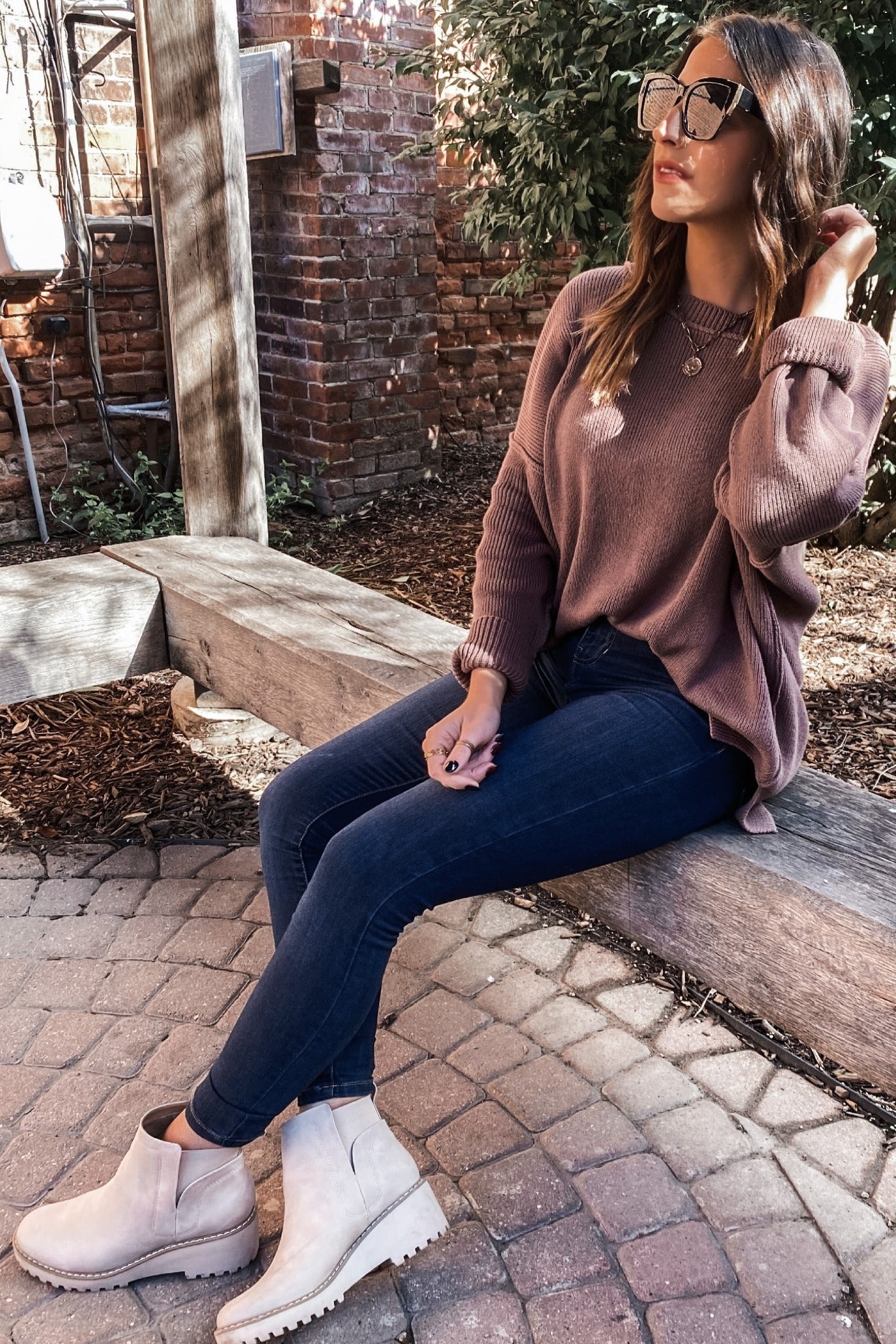 woman wearing a mauve oversized sweater, blue skinny jeans, and beige ankle boots, sitting on a bench outdoors