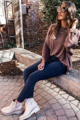woman wearing a mauve oversized sweater, blue skinny jeans, and beige ankle boots, sitting on a bench outdoors