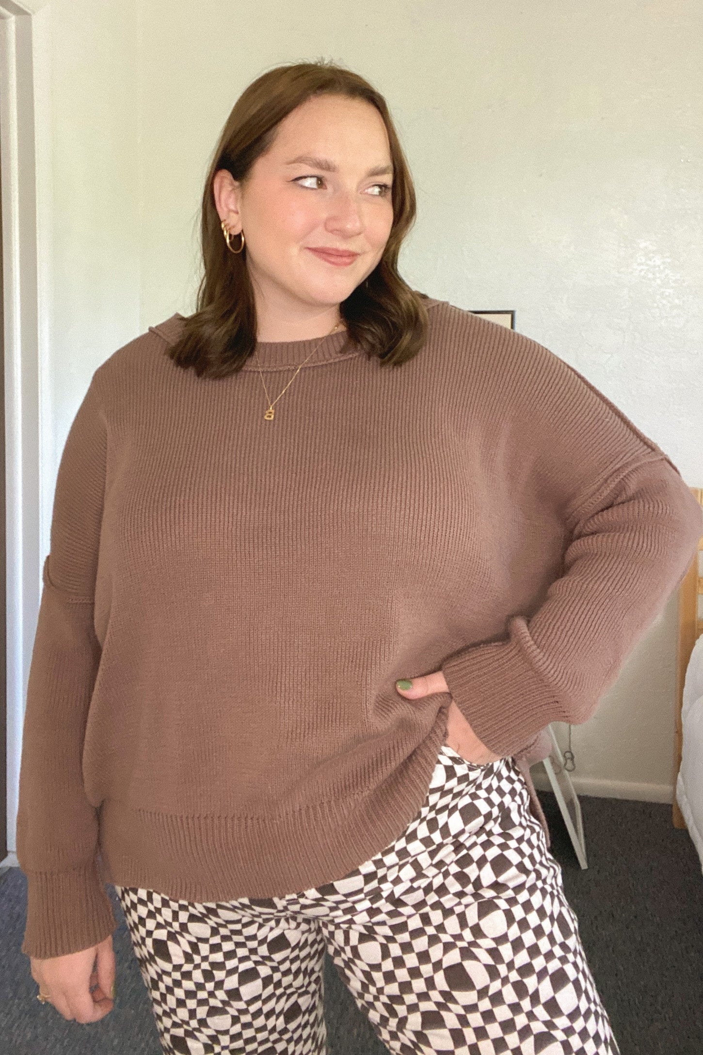 woman wearing a brown oversized knit sweater and black and white patterned pants, smiling slightly with hands on hips