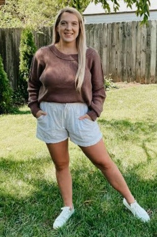 woman wearing a brown long sleeve sweater and white drawstring shorts, standing in a grassy area