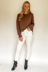 woman wearing a brown oversized sweater and white fitted jeans, standing with hands on hips