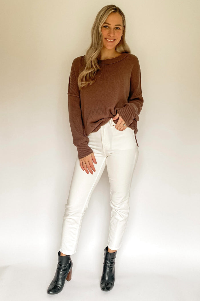 woman wearing a brown oversized sweater and white fitted jeans, standing with hands on hips