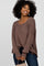 woman wearing a loose brown ribbed sweater and dark jeans, smiling while holding a small handbag