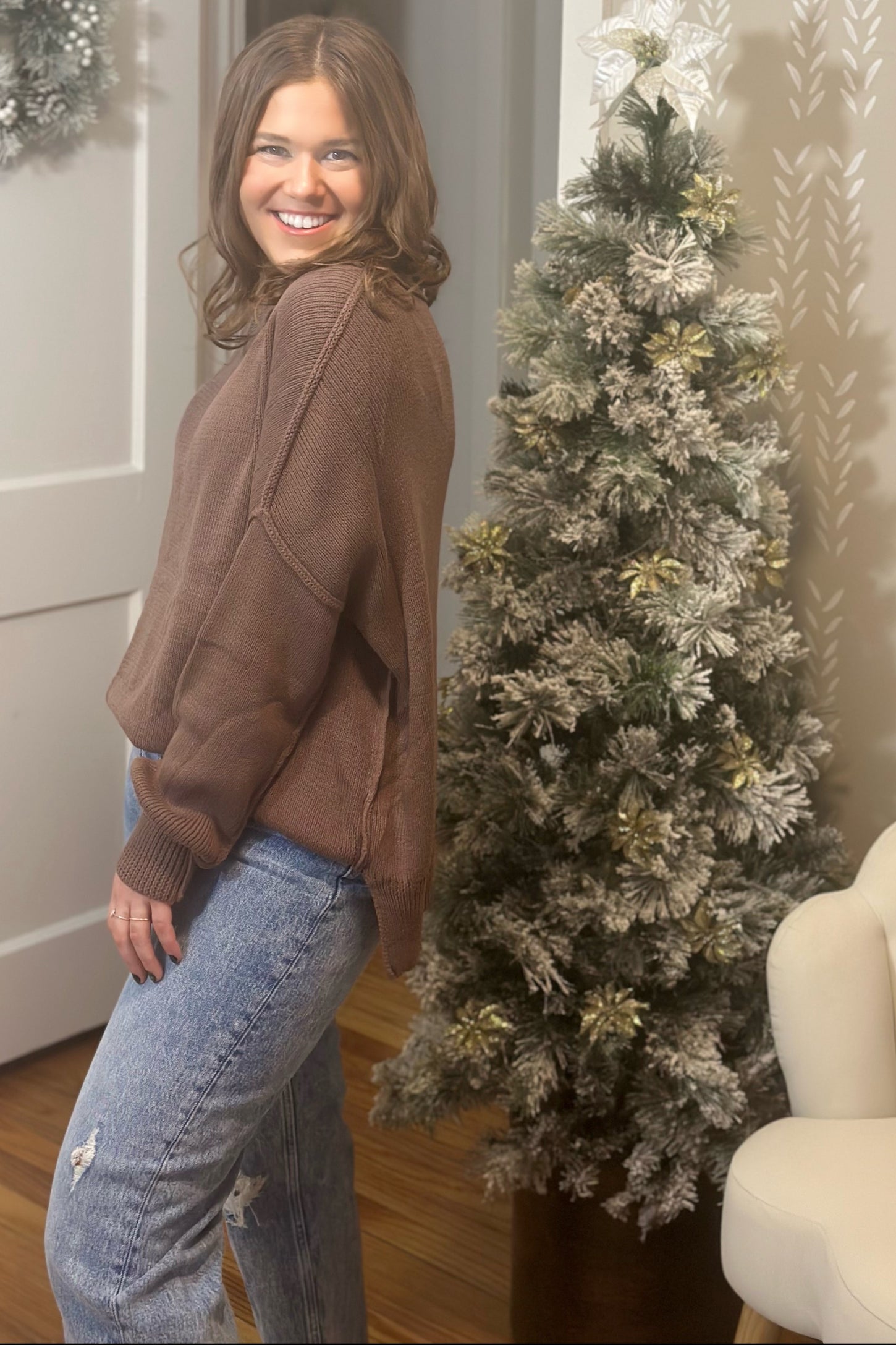woman wearing a brown oversized sweater and high-waisted jeans, standing next to a decorated Christmas tree