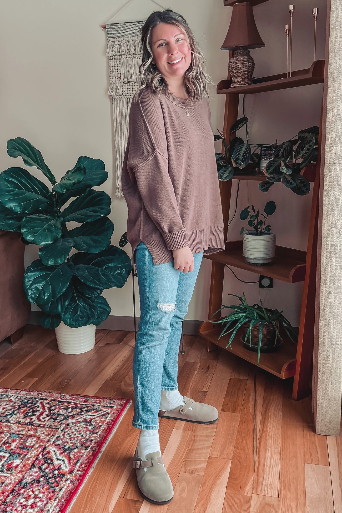 woman wearing a brown oversized sweater, light blue distressed jeans, and tan clogs, standing in a cozy indoor setting