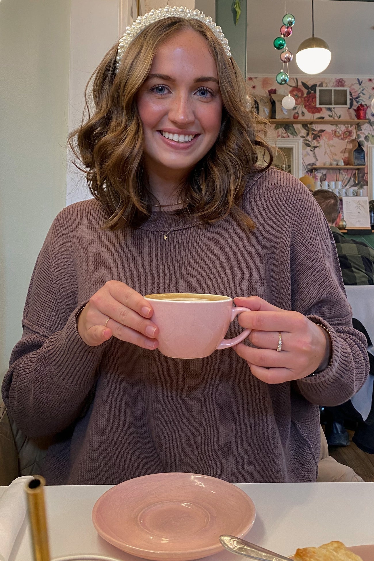 woman wearing a cozy mauve sweater holding a pink cup, sitting at a table with a plate and a decorative background