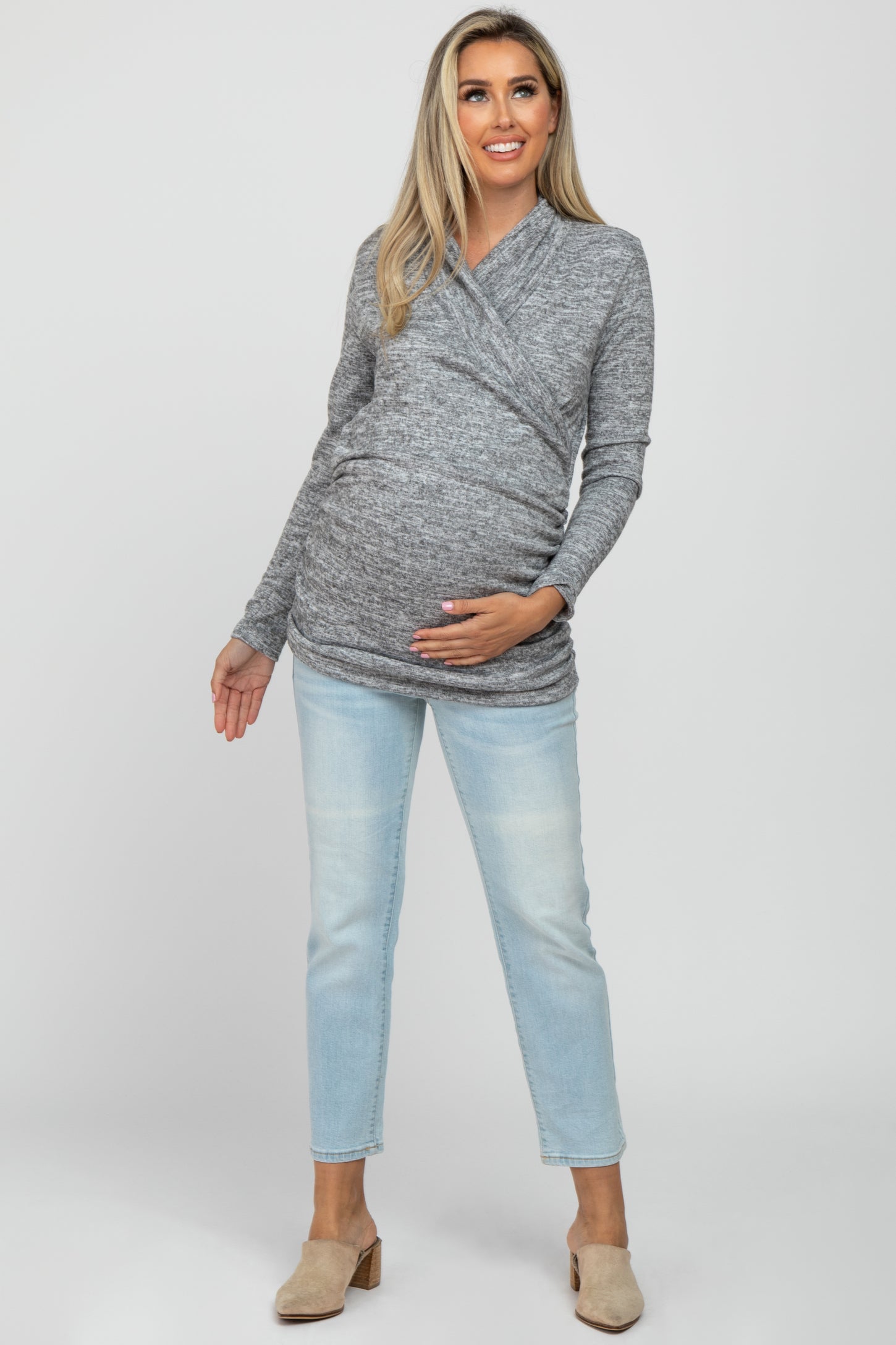 woman wearing a gray wrap maternity top and light blue jeans, holding her baby bump