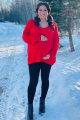woman wearing a red oversized maternity sweater and black leggings, holding her baby bump in a snowy outdoor setting