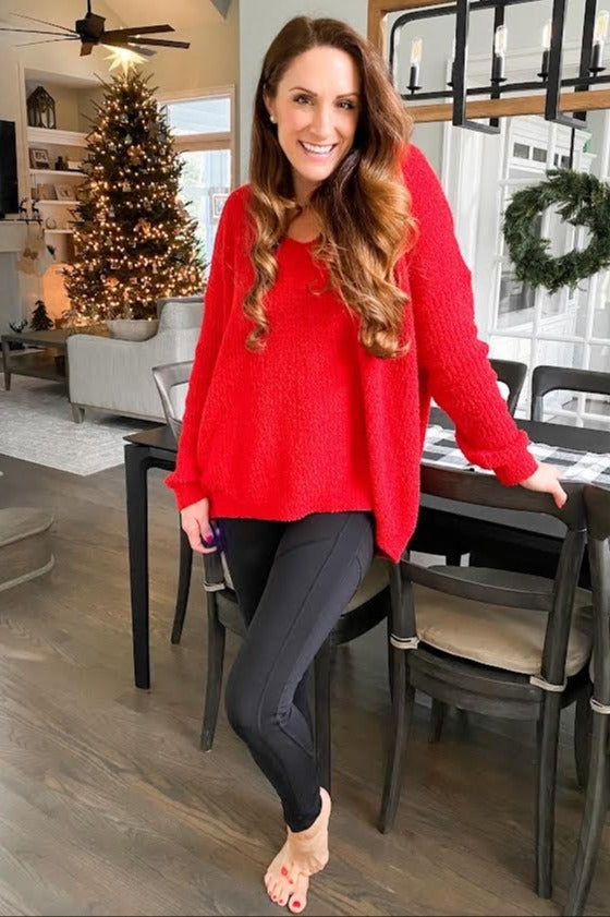 woman wearing a red oversized sweater and black leggings, smiling and posing in a decorated holiday setting