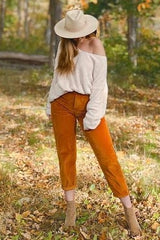 woman wearing a cream off-the-shoulder sweater and orange corduroy pants, standing in a forest setting